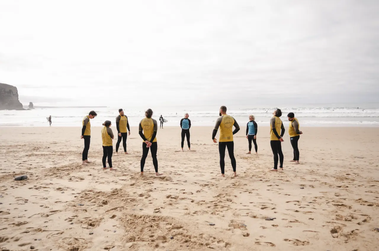 Eine Gruppe in Neoprenanzügen steht im Kreis an einem weiten Sandstrand, im Hintergrund die Brandung.
